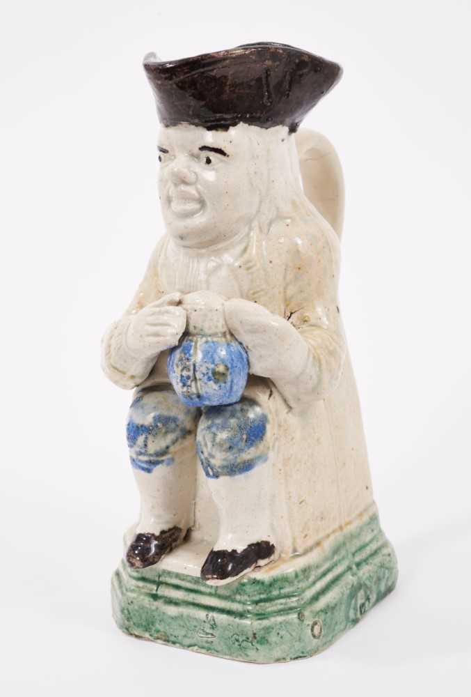 Lot 8 - Creamware Toby jug, circa 1790, shown seated, holding a frothing jug of beer, wearing a cream jacket and blue breeches, on a green square canted base, 16.5cm high