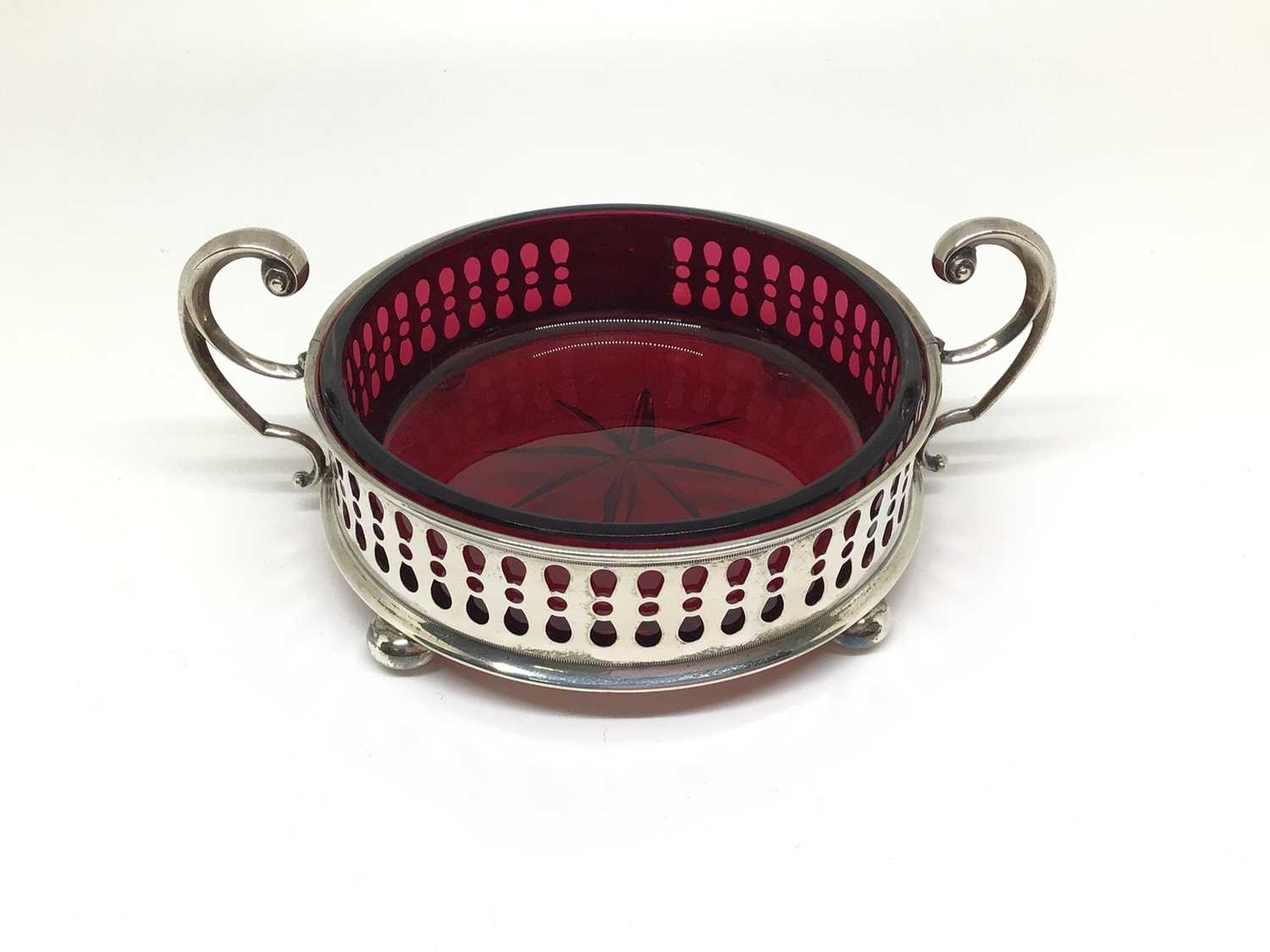 Lot 135 - Edwardian cranberry cut glass dish contained in a silver holder with scroll handles, four bun feet and pierced decoration, 10cm diameter excluding handles, Sheffield 1908 (Watson & Gillott)