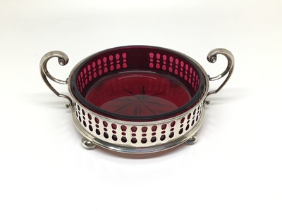 Lot 135 - Edwardian cranberry cut glass dish contained in a silver holder with scroll handles, four bun feet and pierced decoration, 10cm diameter excluding handles, Sheffield 1908 (Watson & Gillott)