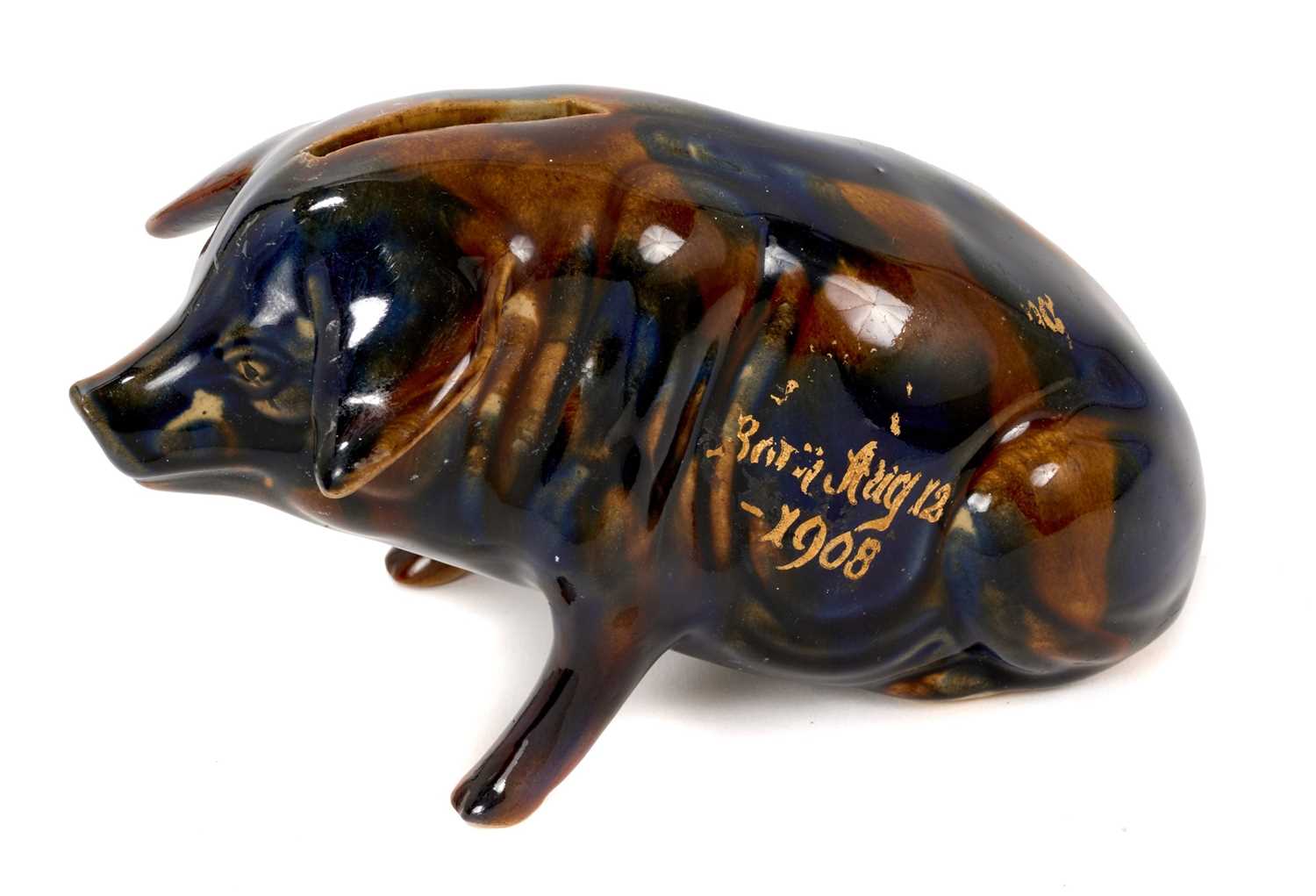Lot 43 - Ewenny pottery model of a pig, the coin slot at the top, decorated with a mottled blue and brown glaze, with partly legible gilt inscription reading 'Born Aug 12-1908', 16.5cm length