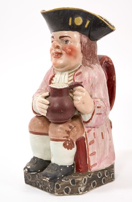 Lot 303 - Early 19th century pearlware glazed Toby jug, wearing a pink jacket, and a yellow and black tricorn hat, on a black spotted base, 25cm high