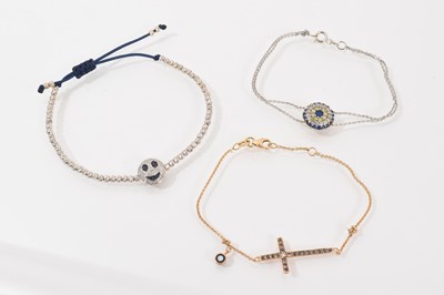 Lot 273 - Three gold diamond and gem set bracelets to include an 18ct white gold diamond and blue sapphire smiley face cord bracelet, 18ct gold diamond and blue sapphire cross pendant bracelet and 14ct wh...