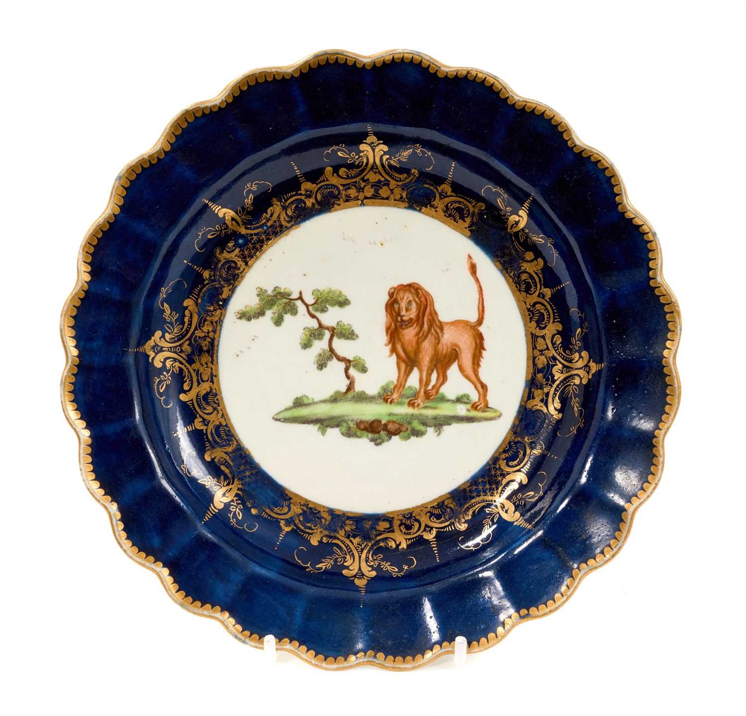 Lot 43 - A Worcester lobed circular plate, circa 1770, painted in the style of Jefferyes Hamett O'Neale (probably by an apprentice) with a lion standing by a tree, on a 'wet blue' ground within a formal...