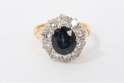 Lot 3432 - Sapphire and diamond cluster ring with an oval mixed cut blue sapphire measuring approximately 9.2mm x 8.2mm x 5mm, surrounded by ten brilliant cut diamonds in claw setting with scroll shoulders on...