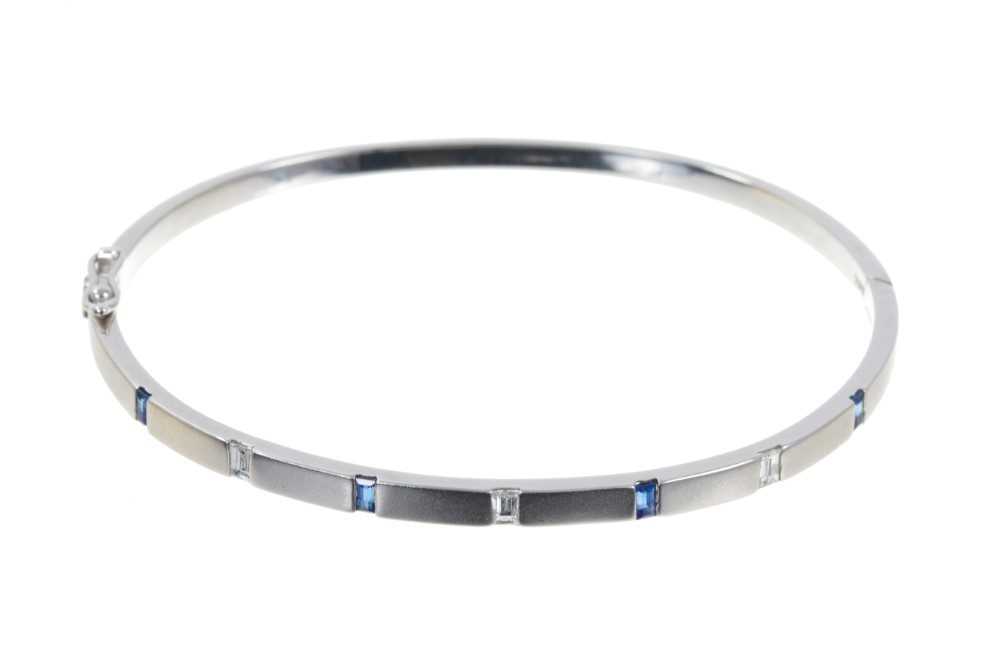 Lot 502 - Contemporary 18ct white gold diamond and blue sapphire hinged bangle with four rectangular step cut blue sapphires interspaced by three baguette cut diamonds with brushed satin finish, stamped 750.
